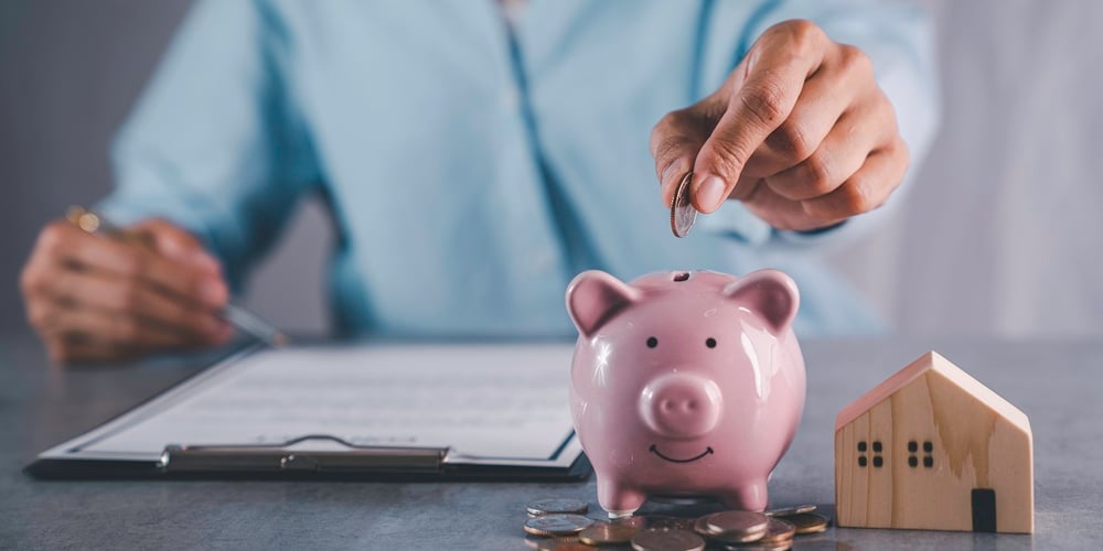 A woman putting a coin in a piggy bank next to small house