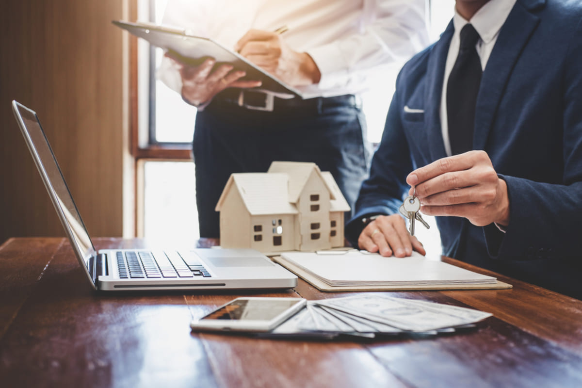 Two businessmen working next to a model house, papers, and keys