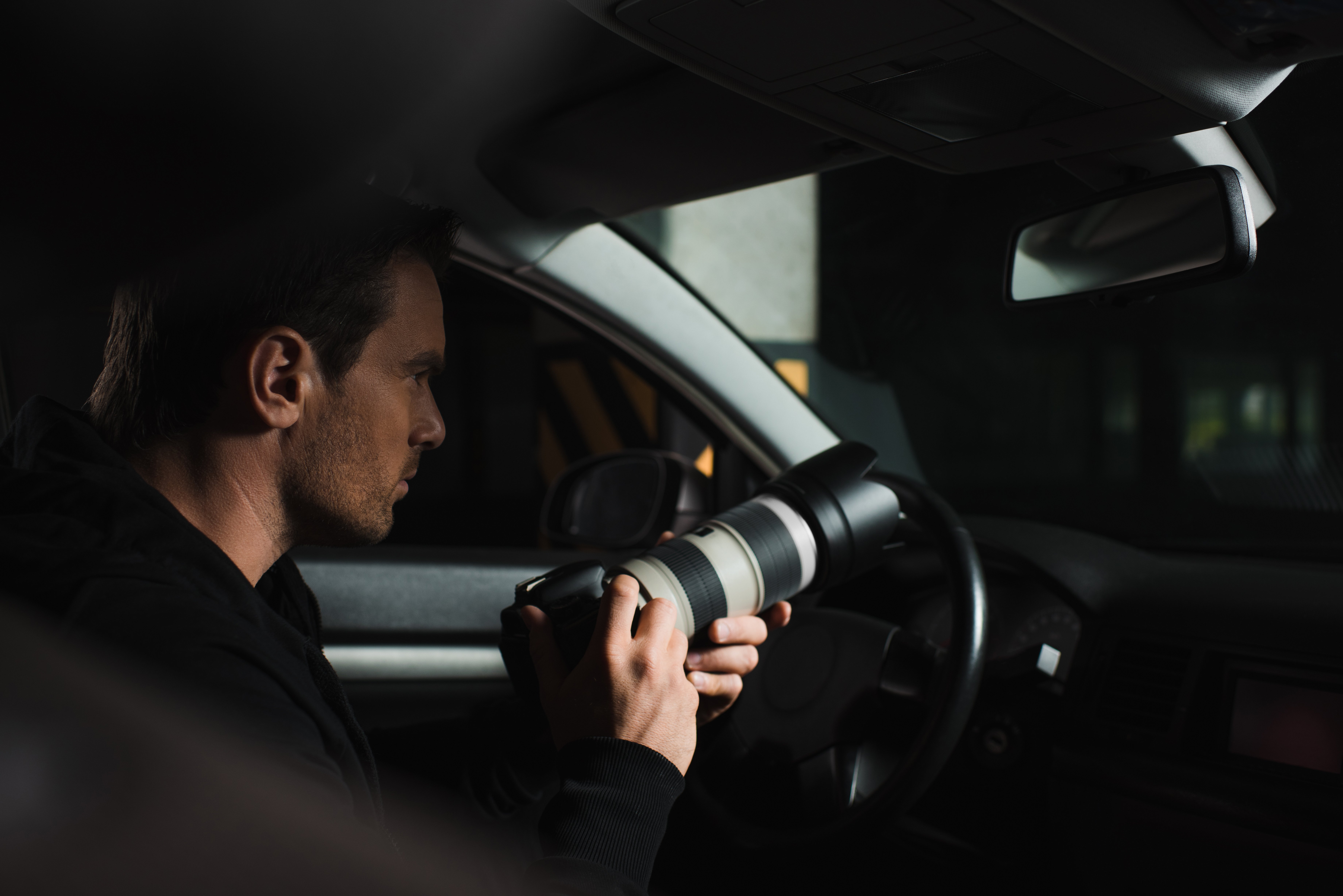 Concentrated male paparazzi doing surveillance by camera with lens from his car