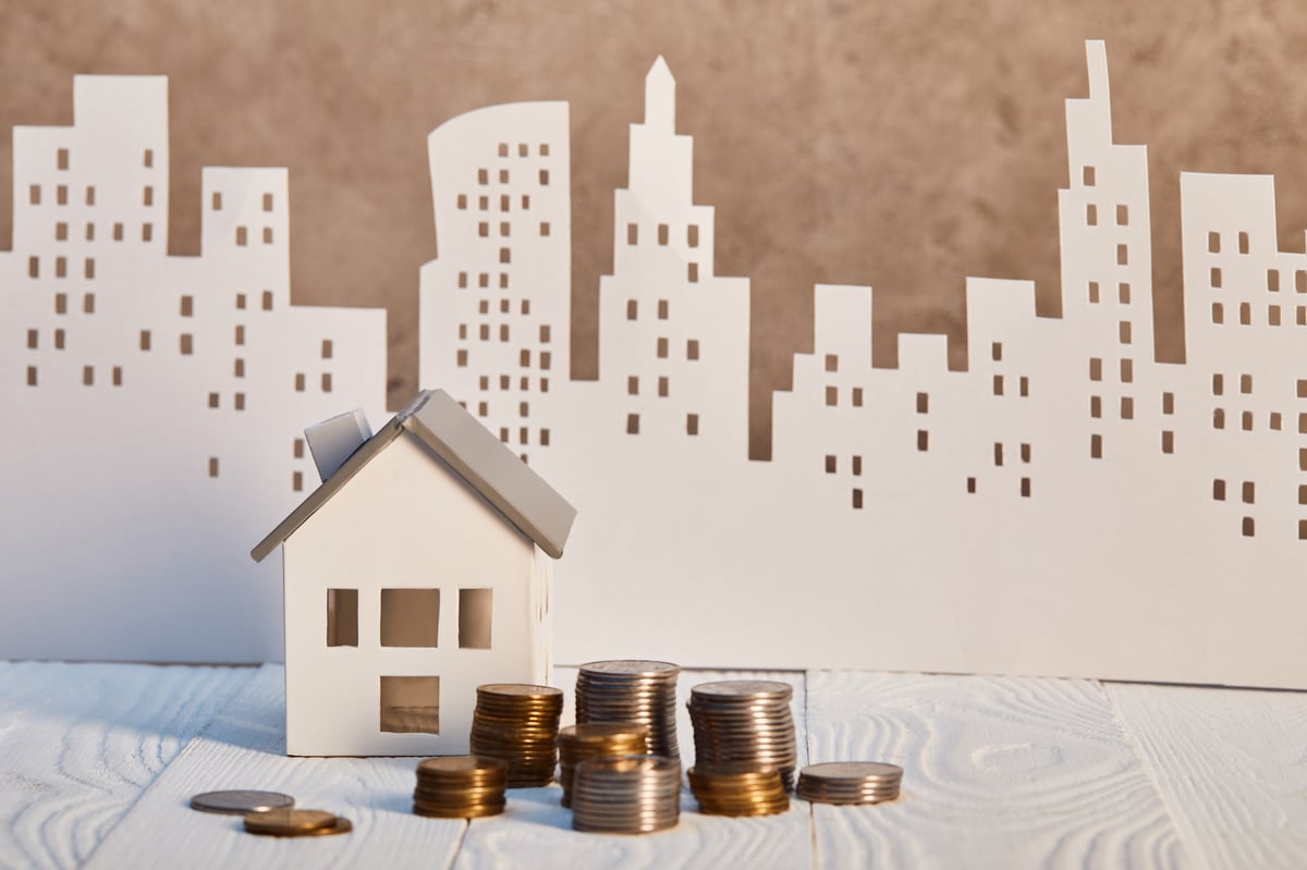 Metal coins and house model on white wooden table near brown textured wall, real estate concept