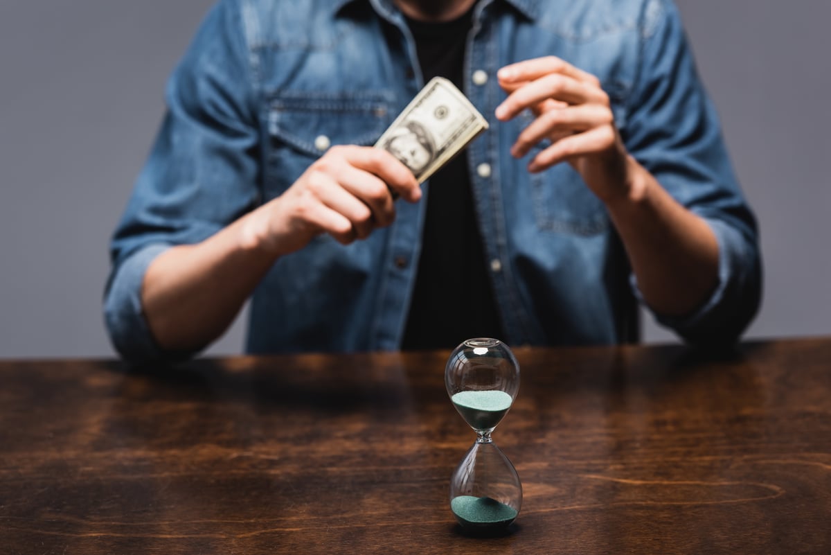 Selective focus of hourglass near man holding dollars while sitting at table isolated on grey, concept of time management