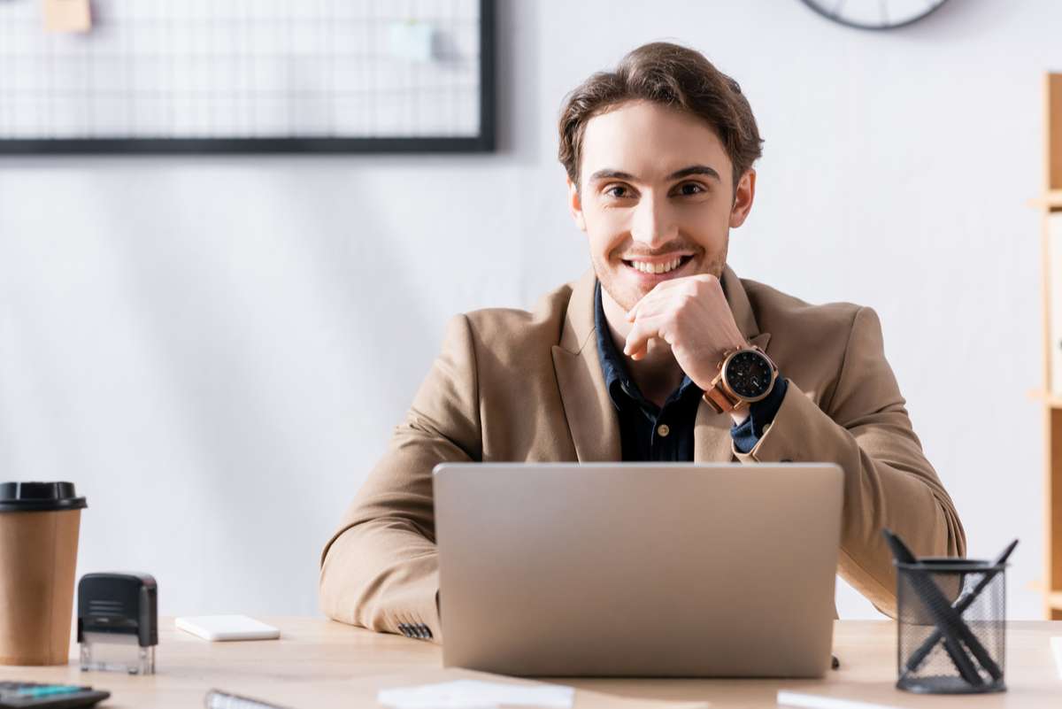 Smiling office worker looking at camera while sitting near laptop at desk in office on blurred background (R) (S)