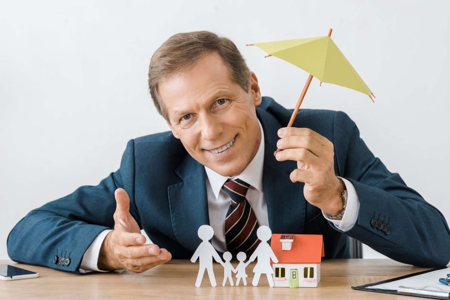 Businessman holding umbrella with paper cut family and house model on wooden table