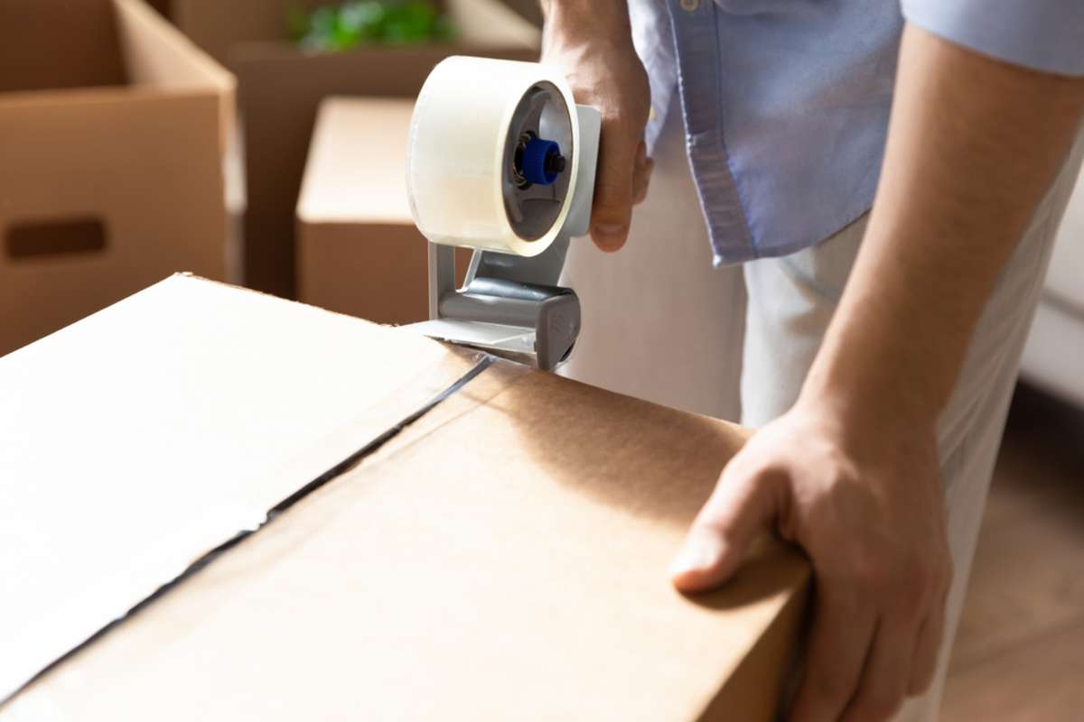 Close up young man packing stuff in cardboard box, using adhesive scotch indoors