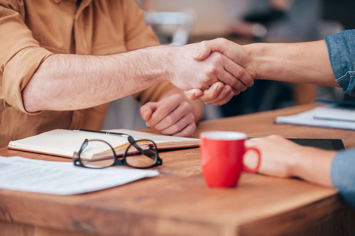 Close-up of two men shaking hands while sitting at the wooden desk-1