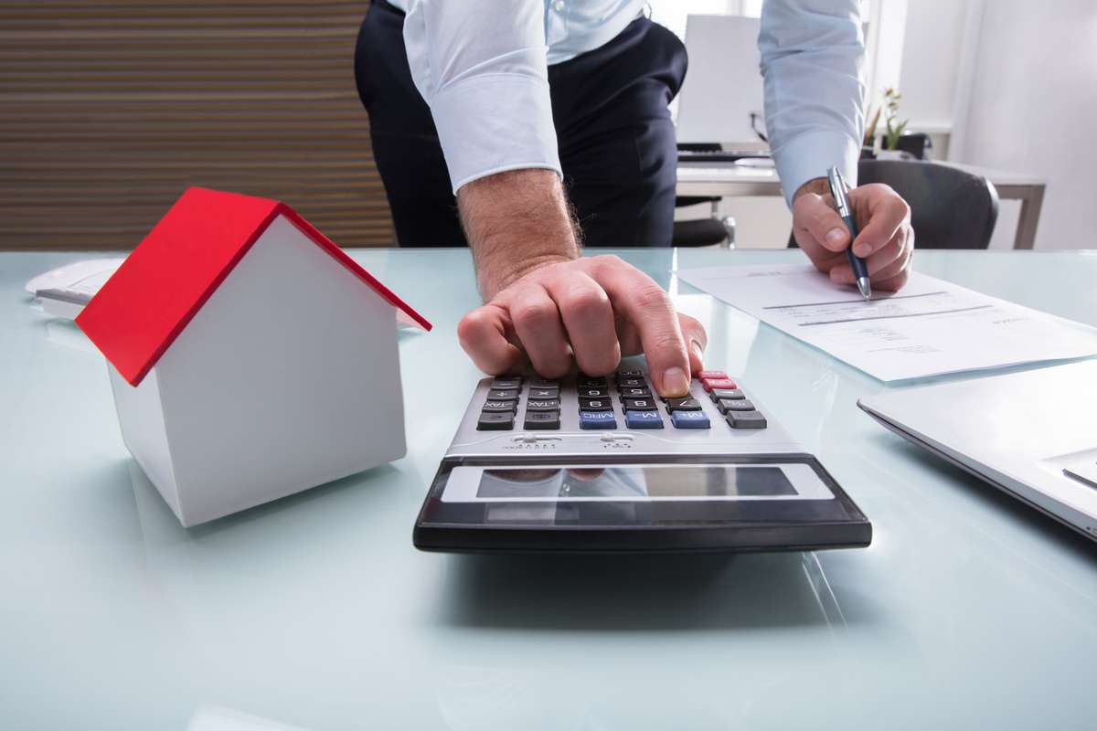 Human Hand Calculating Bill Using Calculator With House Model On Desk