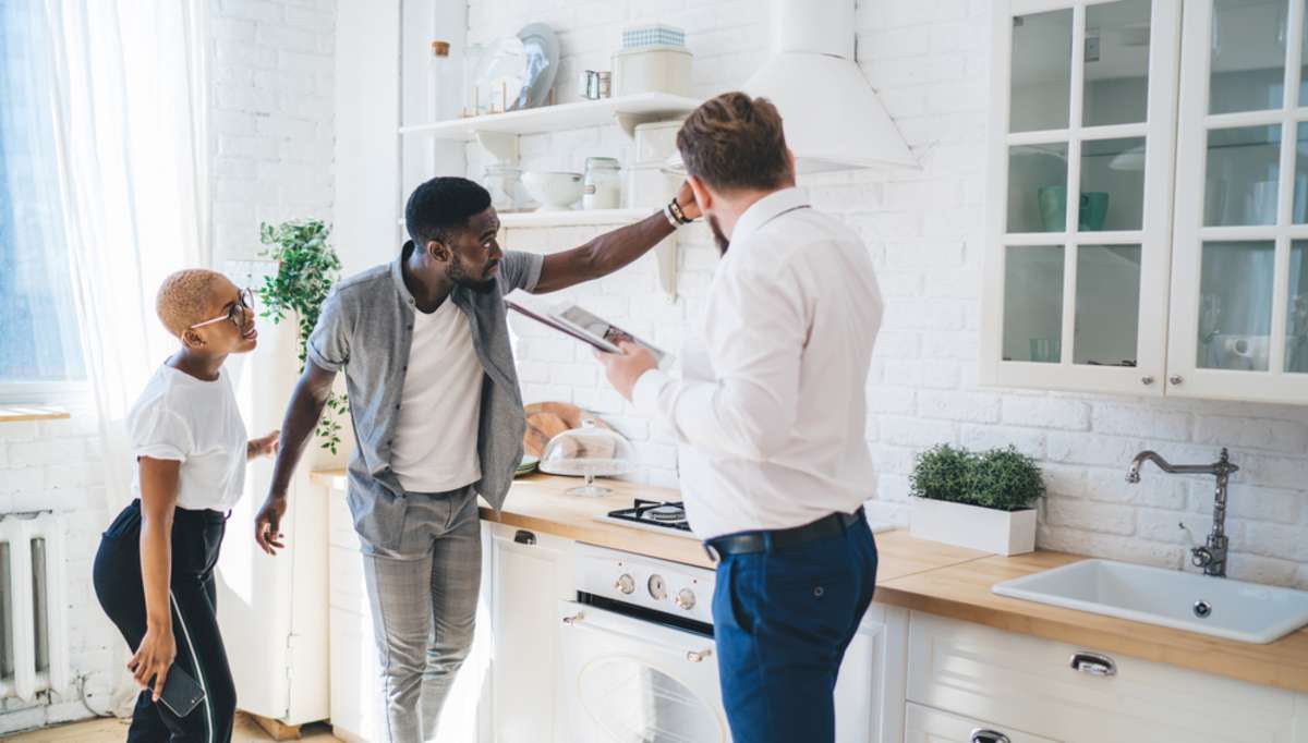 Side view of bearded chubby professional man in formal suit with clipboard inspecting hood fan together with thoughtful ethnic couple while standing in sunny modern apartment