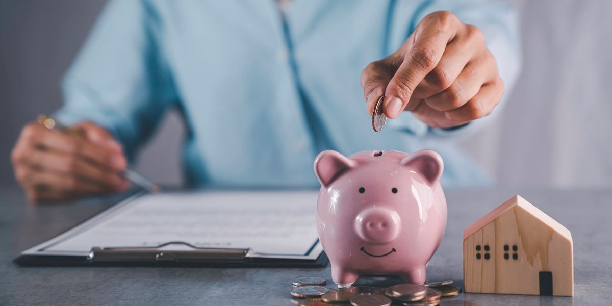 A woman putting a coin in a piggy bank next to small house